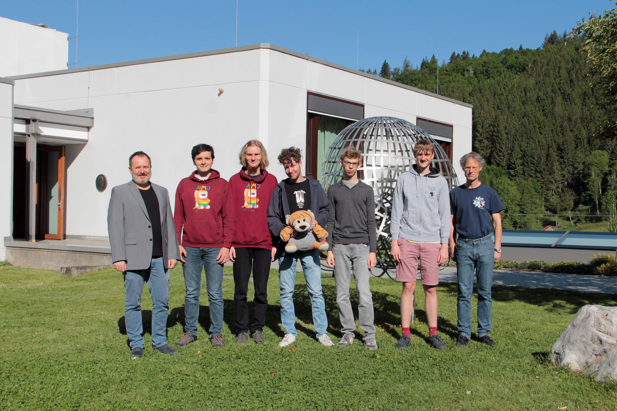 Das Team der IMO 2025-zwei Teilnehmer in rostroten Pullovern mit MathemaTigerin-Aufdruck, außen die beiden Delegationsleiter. 