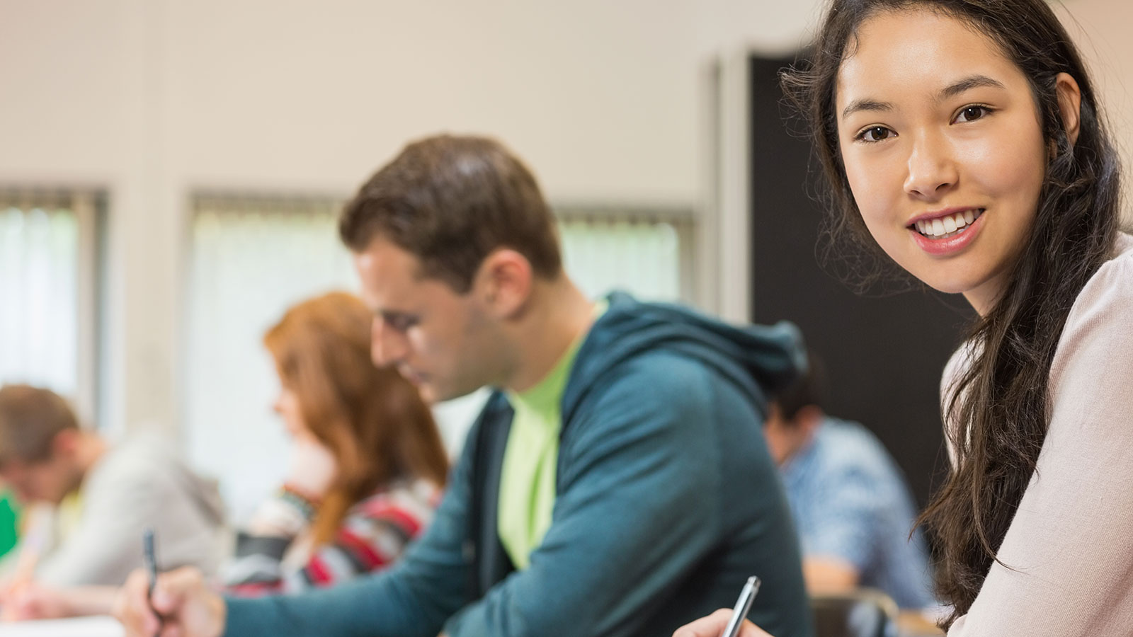 Junge Schülerin sitzt mit anderen Schülern in einem Klassenzimmer und lächelt in die Kamera.