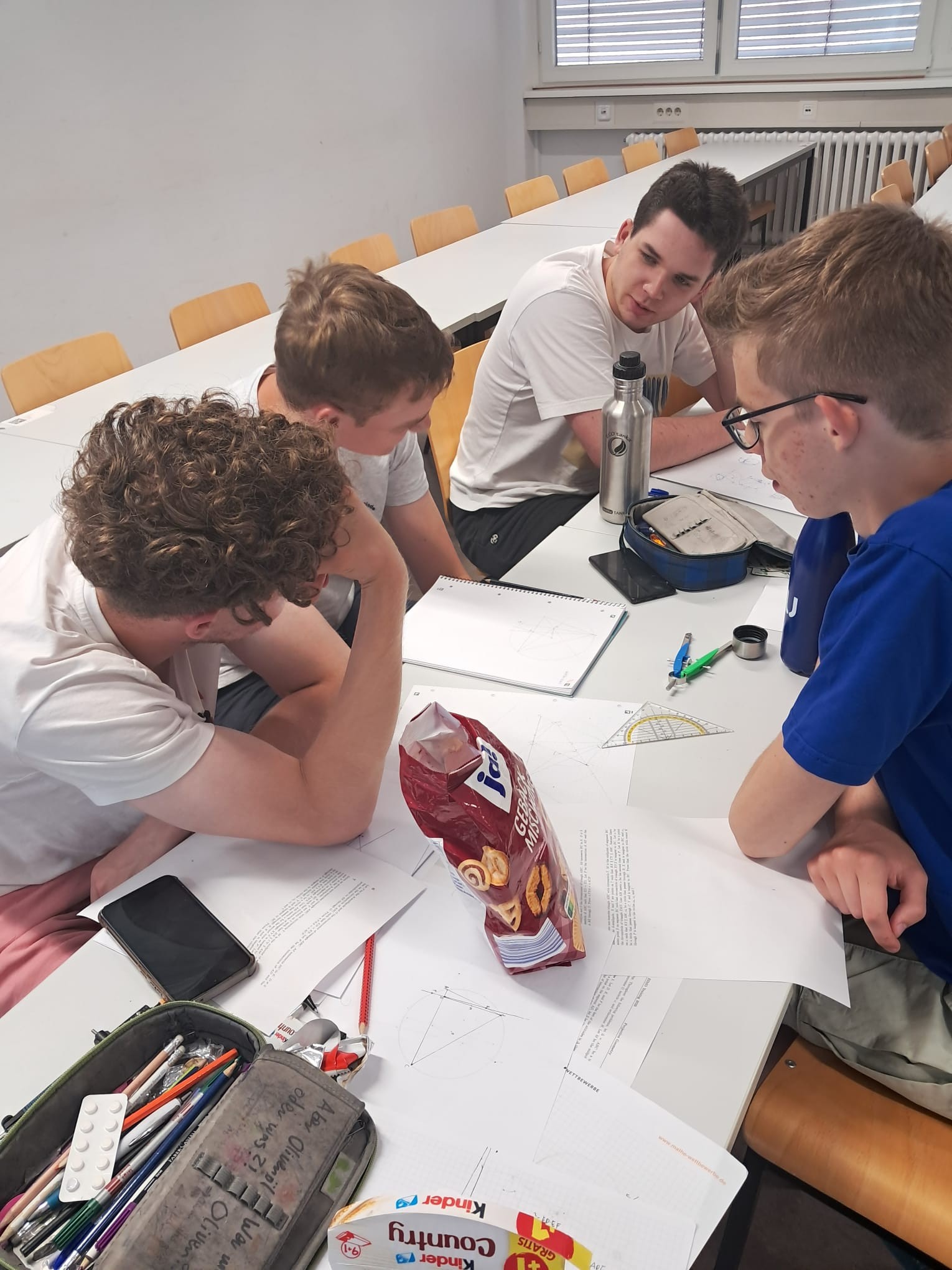 Vier Teilnehmende über einen Tisch gebeugt, Stifte, Papier, Geodreiecke und Süßigkeiten sind auf dem Tisch verteilt.