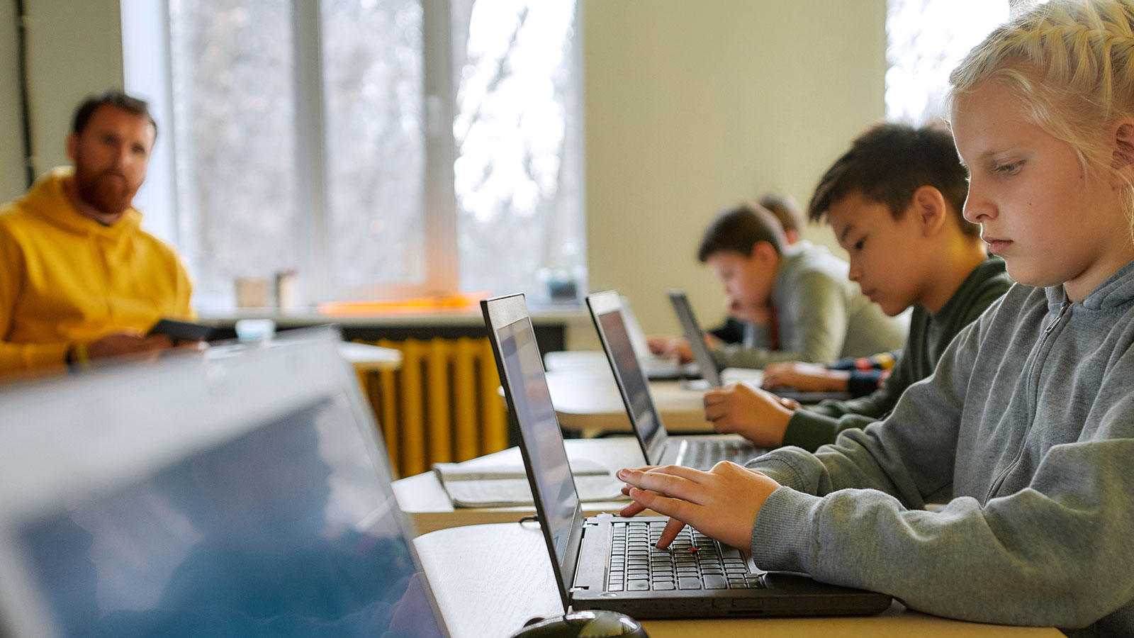 Junge Schülerinnen und Schüler sitzen mit einem Lehrer in einem Klassenraum und arbeiten an Laptops.