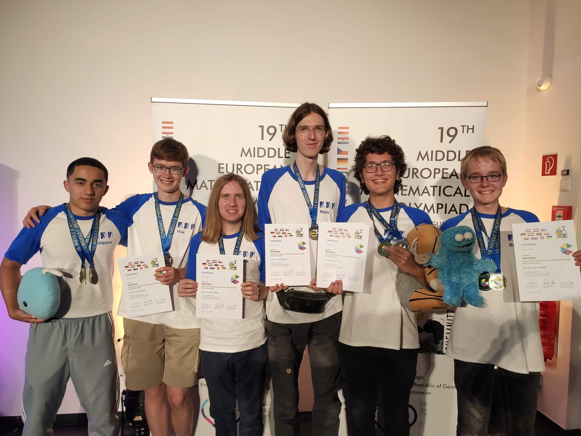 Gruppenfoto: Die deutsche Delegation präsentiert stolz ihre Medaillen und Urkunden, im Hintergrund zwei Banner mit den Förderern und mit dem Logo der MeMo