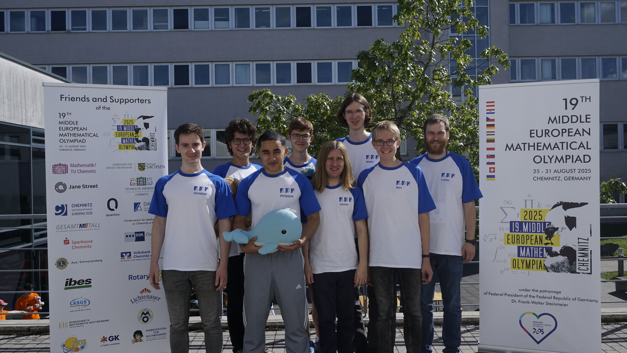 Gruppenfoto: Die deutsche Delegation auf dem Gelände der TU Chemnitz, links ein Banner mit den Förderern, rechts ein Banner mit dem Logo der MeMo