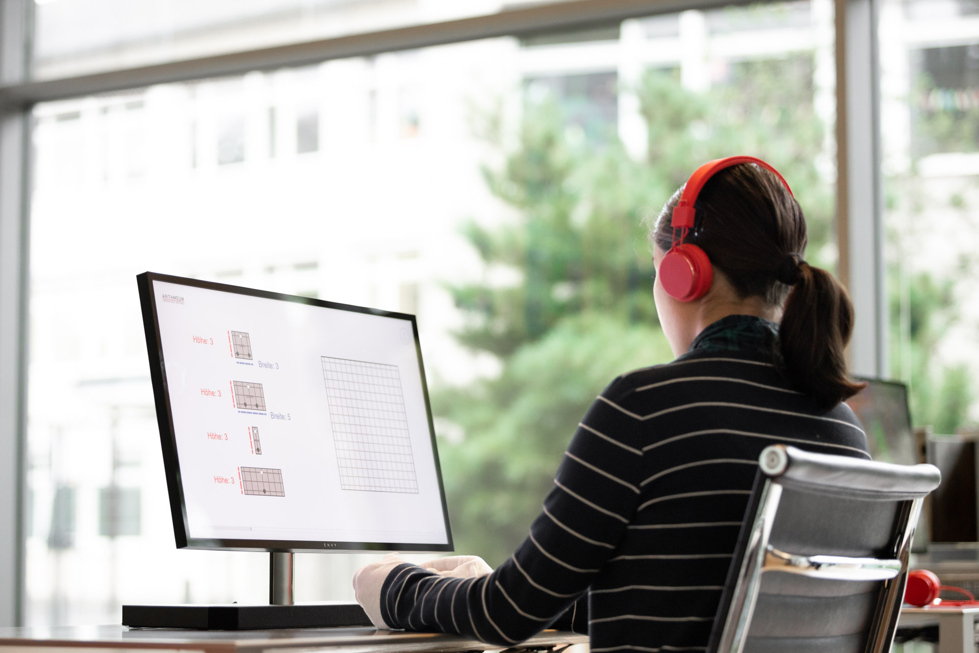 Ein Mädchen mit roten Kopfhörern vor einem PC-Bildschirm, von hinten fotografiert. 