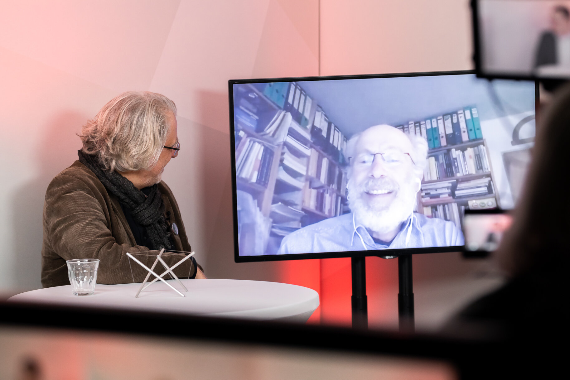 Ein Gast auf der Bühne schaut auf den Bildschirm, wo ein weiterer Gast digital zugeschaltet redet und lacht.