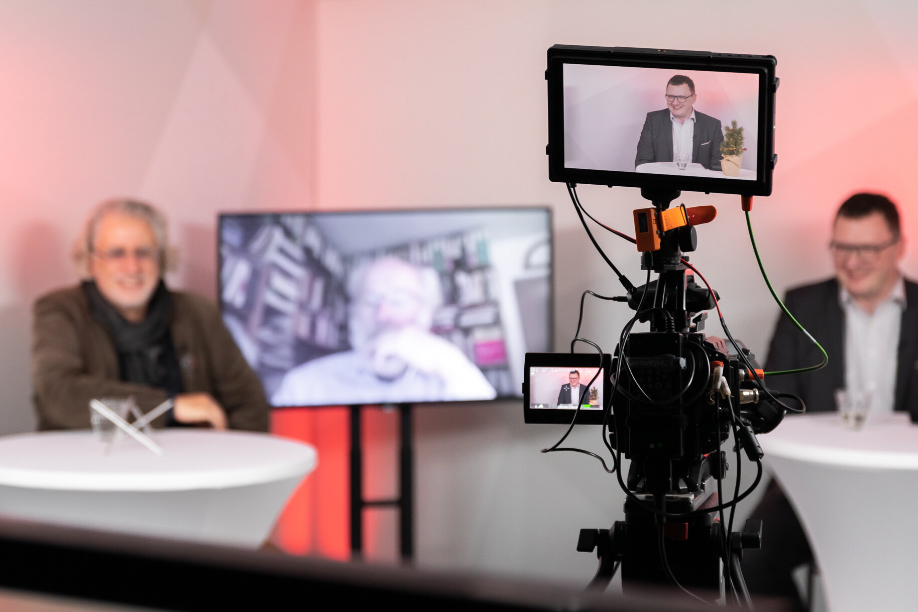 Vorne eine Kamera im Fokus. Unscharf im Hintergrund ein Panel aus drei Personen, davon eine digital zugeschaltet.