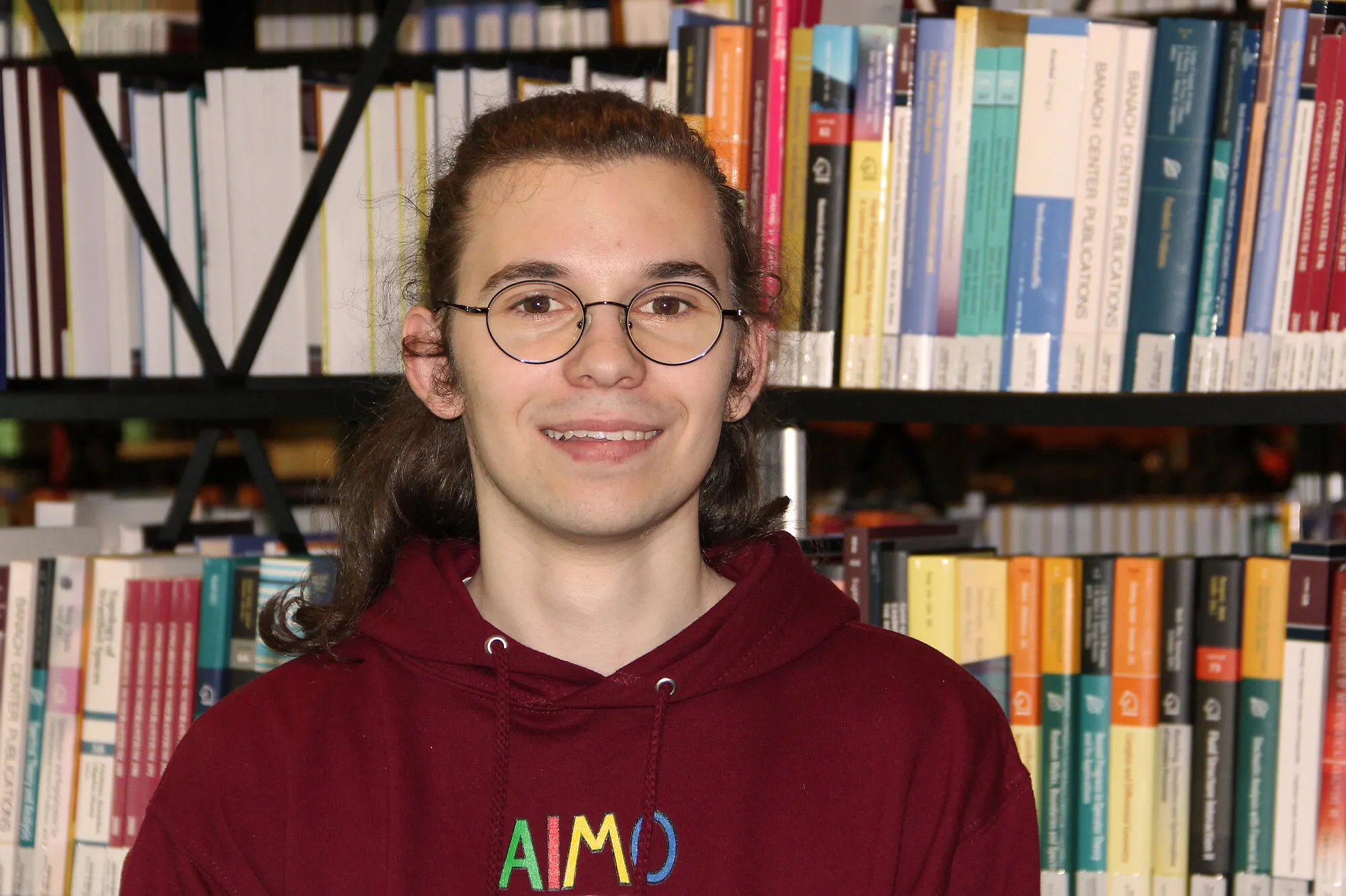 Christian Kraftsik mit IMO 2024-Pullover vor einem Bücherregal. 