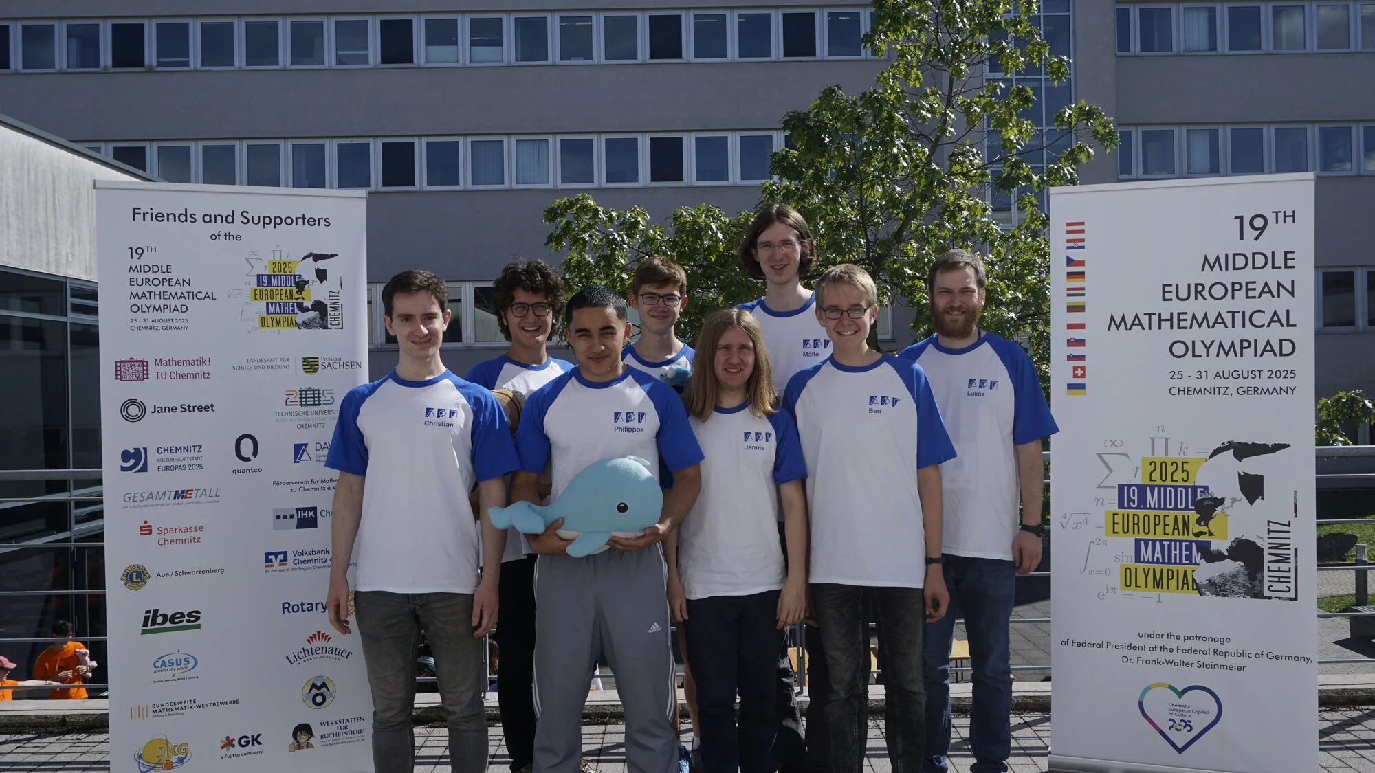 Gruppenfoto: Die deutsche Delegation auf dem Gelände der TU Chemnitz, links ein Banner mit den Förderern, rechts ein Banner mit dem Logo der MeMo